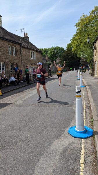 Emily Green runs towards the finish of the Eyam Half Marrathon, her arms raised in celebration. Another runner is in the foreground.