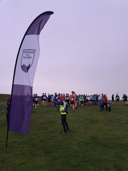 A pre-race briefing. Runners are in the background, listening to a figure in hi-vis in the middleground. In the foreground, to the left, is a "Durham Fell Runners" banner.